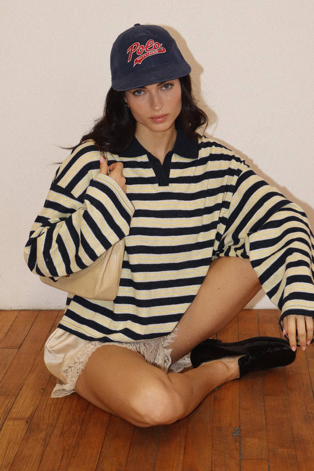Model seated on the floor in the Stripe Polo Top in black and cream stripes with a navy cap